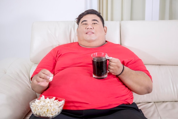 Hombre gordo viendo la televisión mientras sostiene un vaso de cola