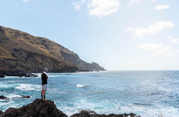 Hombre con gorra de pie en las rocas mirando el paisaje marino en un día claro