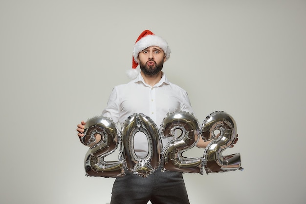 Foto un hombre con un gorro de papá noel de terciopelo rojo sostiene globos plateados con la forma de 2022. un tipo sorprendido con barba en una fiesta de año nuevo.