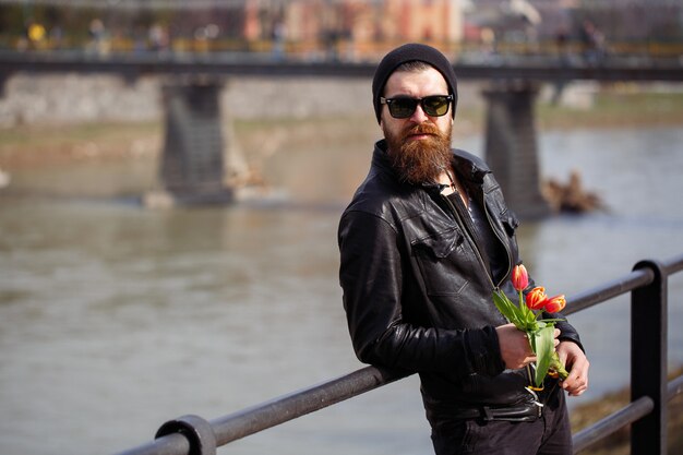 Un hombre con una gran barba tupida con un gorro y gafas de sol con una chaqueta de cuero y pantalones oscuros se encuentra con tulipanes rojos sobre el fondo de un puente peatonal y un río