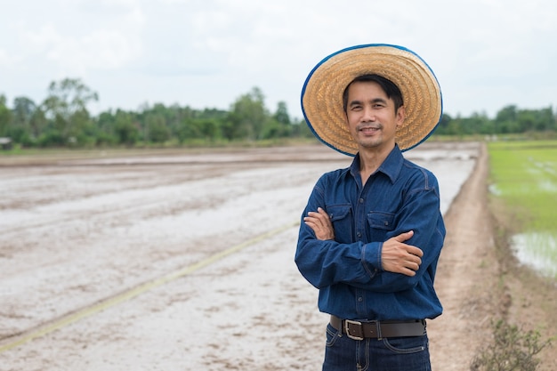 El hombre del granjero asiático usa la camisa azul y el soporte del sombrero con el brazo cruzado en la granja de arroz verde.