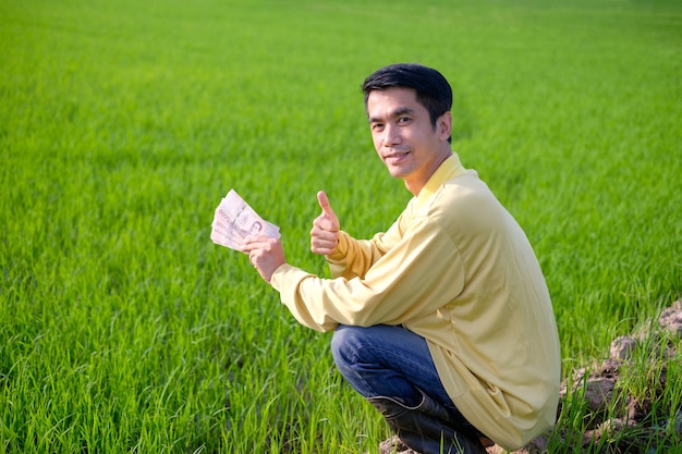 Hombre de granjero asiático viste camisa amarilla sentado y sosteniendo dinero de billetes tailandeses en la granja de arroz verde.