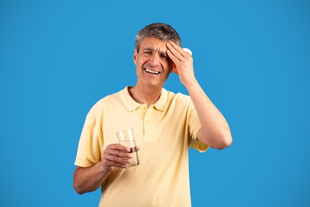 Hombre gris que sufre de dolor de cabeza tomando pastillas sobre fondo azul.