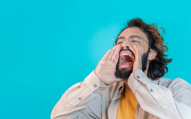 Hombre gritando fuerte llamando la atención con las manos como un megáfono aislado en el fondo azul del estudio