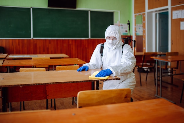 Un hombre del grupo de desinfección limpia el pupitre de la escuela con un trapo amarillo. Un trabajador profesional esteriliza el aula para evitar la propagación del covid-19. Concepto de salud de alumnos y estudiantes.