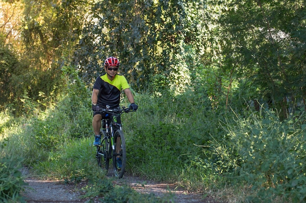 Hombre con guante de casco para la seguridad que monta una bicicleta en el camino rural a lo largo de un bosque