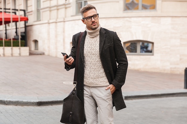 Hombre guapo con un abrigo caminando al aire libre, sosteniendo el teléfono móvil