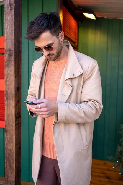 Hombre guapo con abrigo usando teléfono inteligente cerca de una casa de madera.