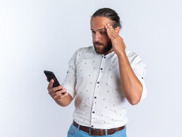 Hombre guapo adulto confundido con gafas manteniendo la mano en la cabeza sosteniendo y mirando el teléfono móvil aislado en la pared blanca
