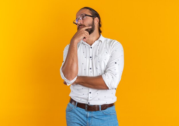 Hombre guapo adulto pensativo con gafas manteniendo la mano en la barbilla mirando al lado aislado en la pared naranja con espacio de copia