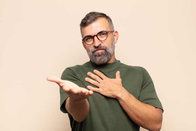 Hombre guapo adulto que se siente feliz y enamorado, sonriendo con una mano al lado del corazón y la otra estirada al frente