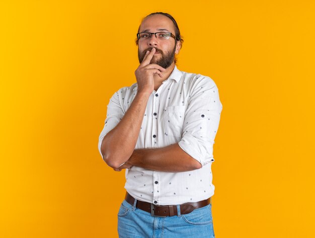 Hombre guapo adulto reflexivo con gafas manteniendo los dedos en los labios mirando a la cámara aislada en la pared naranja con espacio de copia