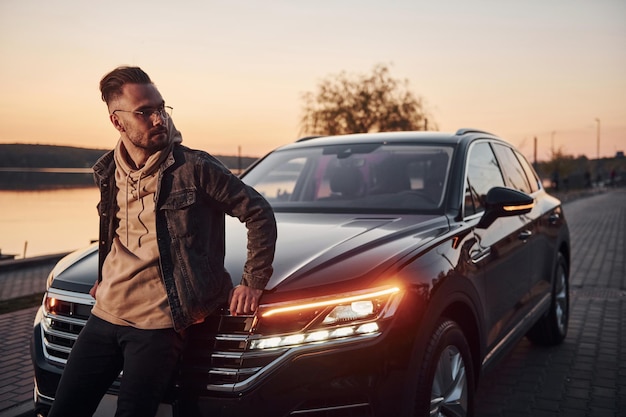 Foto hombre guapo sin afeitar con ropa de moda apoyado en su auto negro