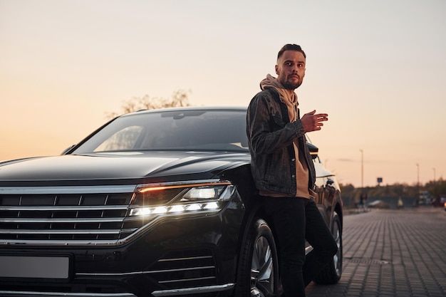 Foto hombre guapo sin afeitar con ropa de moda parado cerca de su auto negro y fumando