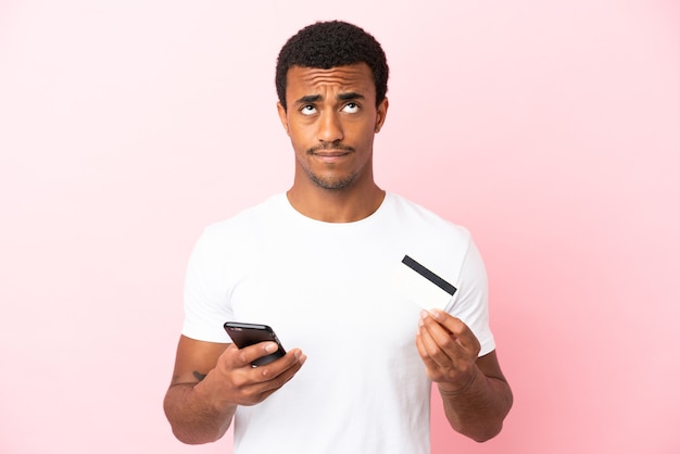 Hombre guapo afroamericano sobre fondo rosa aislado comprando con el móvil con una tarjeta de crédito mientras piensa