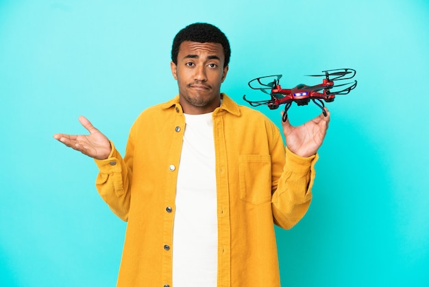 Hombre guapo afroamericano sosteniendo un avión no tripulado sobre fondo azul aislado teniendo dudas mientras levanta las manos