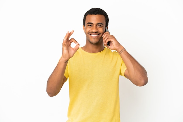 Hombre guapo afroamericano mediante teléfono móvil sobre fondo blanco aislado mostrando el signo de ok con los dedos