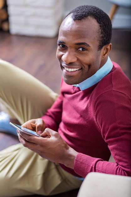 Hombre guapo agradable alegre sonriendo y mirando directamente mientras usa su dispositivo móvil