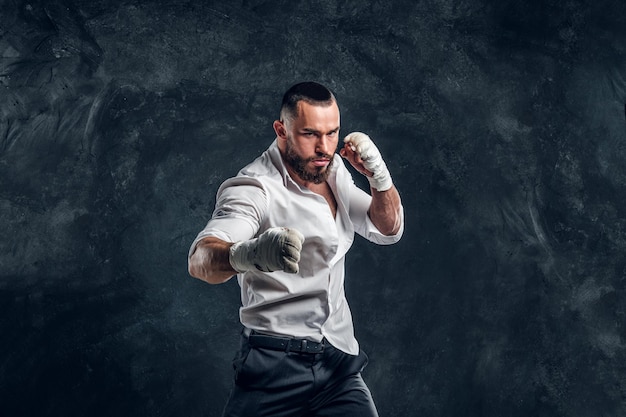 Un hombre guapo y agresivo con camisa blanca está demostrando su golpe en un estudio fotográfico oscuro.