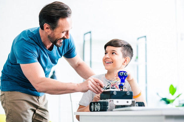 Hombre guapo alegre ayudando a su hijo mientras prueba un nuevo dispositivo robótico en casa