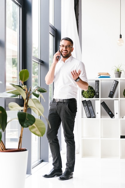 Foto hombre guapo alegre en ropa formal que tiene una agradable conversación móvil, mientras está de pie cerca de una ventana grande en la oficina