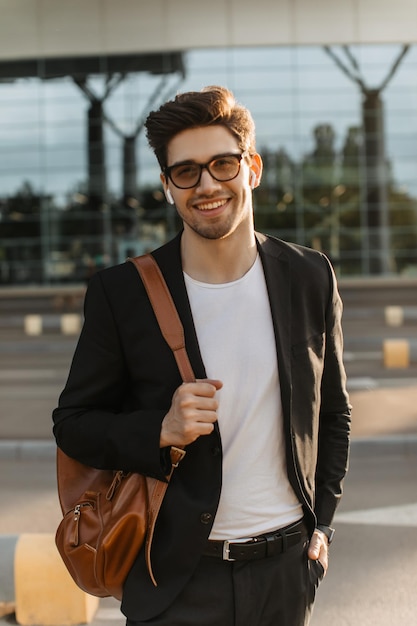 Un hombre guapo con anteojos sonríe sinceramente afuera Retrato de un chico moreno con una chaqueta negra camiseta blanca sosteniendo una mochila cerca del aeropuerto