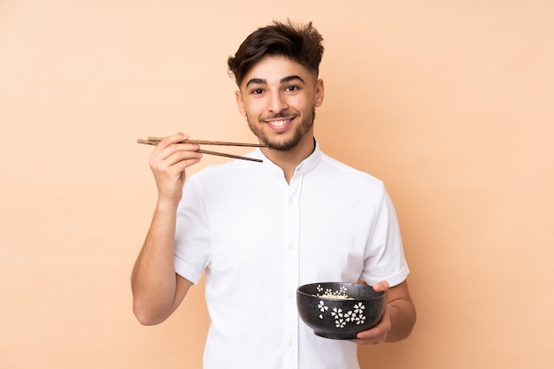 Hombre guapo árabe en la pared de color beige con un tazón de fideos con palillos