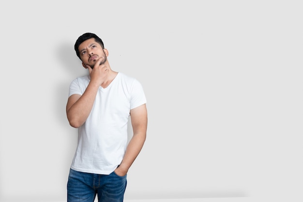 Hombre guapo asiático en camiseta blanca que tiene dudas mientras mira hacia arriba sobre fondo blanco aislado