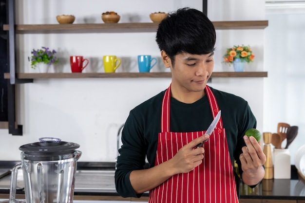 Un hombre guapo asiático está mirando la fruta de limón en su mano y la otra mano sosteniendo un cuchillo en la cocina moderna.