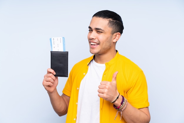 Hombre guapo asiático en pared azul en vacaciones sosteniendo un pasaporte y avión con el pulgar hacia arriba