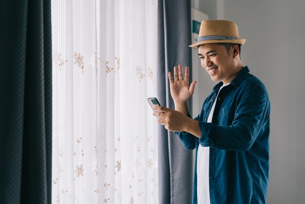 Hombre guapo asiático pasar la mañana en la casa y tener video chat a través del teléfono smar junto a la ventana felizmente.