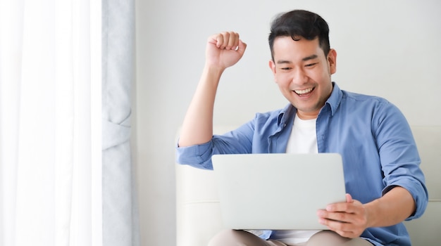 Hombre guapo asiático que trabaja con la computadora portátil en la sala de estar feliz y cara de sonrisa