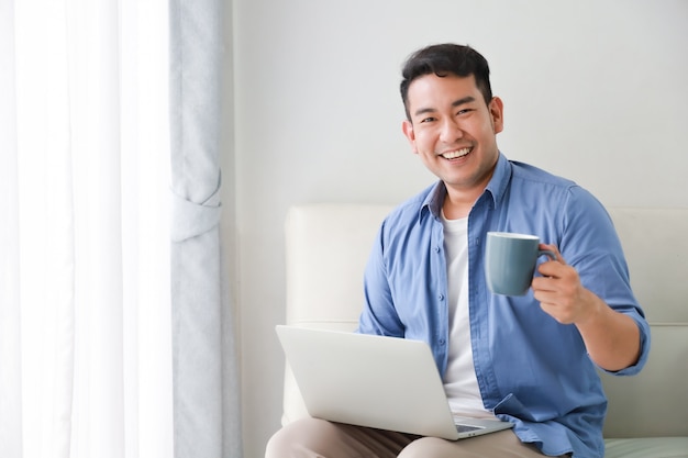 Hombre guapo asiático trabajando con la computadora portátil y tomar café en la sala de estar feliz y cara de sonrisa