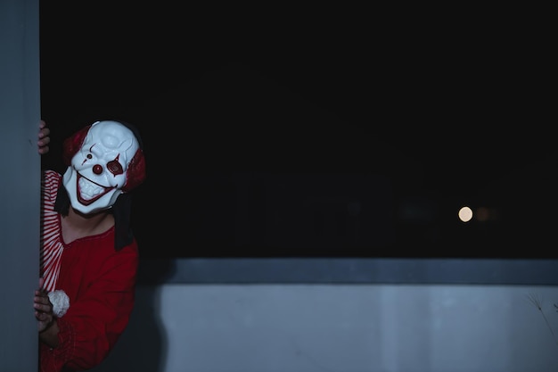 Hombre guapo asiático usa máscara de payaso con arma en la escena nocturna Concepto del festival de Halloween Foto aterradora de un asesino en tela naranja Personaje de payaso malvado