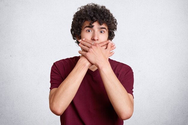 Hombre guapo asustado con el cabello crujiente, contras boca mientras intenta dejar de hablar, usa ropa casual, posa en blanco. Adolescente asustada cotillea con amigos. ¡Estoy callado!