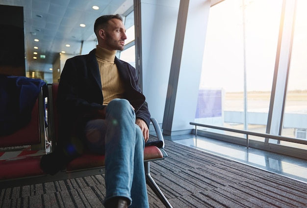 Hombre guapo y atractivo, hombre de negocios confiado en viaje de negocios, sentado en la sala de embarque, admirando la vista desde las ventanas panorámicas del aeropuerto internacional hasta la pista con aviones