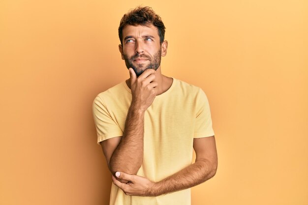 Hombre guapo con barba con camiseta amarilla informal sobre fondo amarillo con la mano en la barbilla pensando en la expresión pensativa de la pregunta sonriendo con el concepto de duda en la cara pensativa