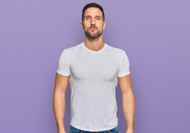 Hombre guapo con barba con camiseta blanca casual relajado con expresión seria en la cara simple y natural mirando a la cámara