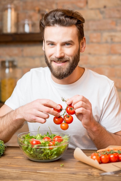 Foto hombre guapo con barba en camiseta blanca haciendo ensalada con tomate y pimiento en la cocina. concepto de comida sana y vegana