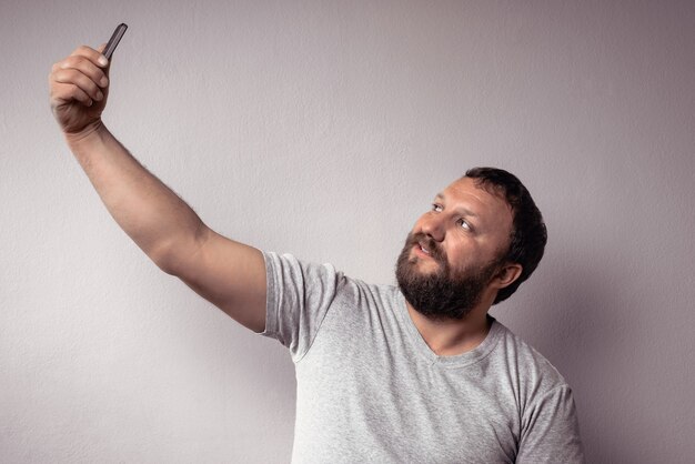 Hombre guapo con barba en camiseta gris mirando el teléfono inteligente y tomando selfie contra la pared gris.