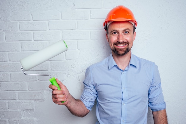 Hombre guapo con barba y un casco casco sobre un fondo de una pared de ladrillo blanco sostiene un cepillo de rodillo.