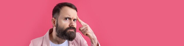 Un hombre guapo con barba en una chaqueta rosa está pensando en un fondo rojo aislado