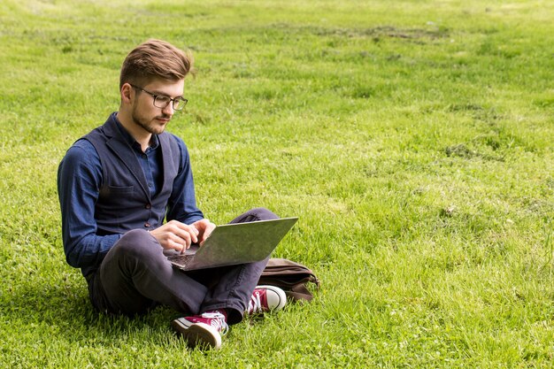 Hombre guapo con barba está sentado en un césped y trabaja en una computadora portátil