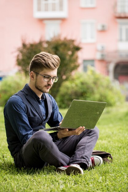 Hombre guapo con barba está sentado en un césped y trabaja en una computadora portátil