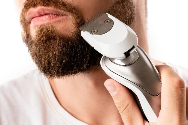 Hombre guapo con barba está utilizando una recortadora eléctrica sobre fondo blanco.