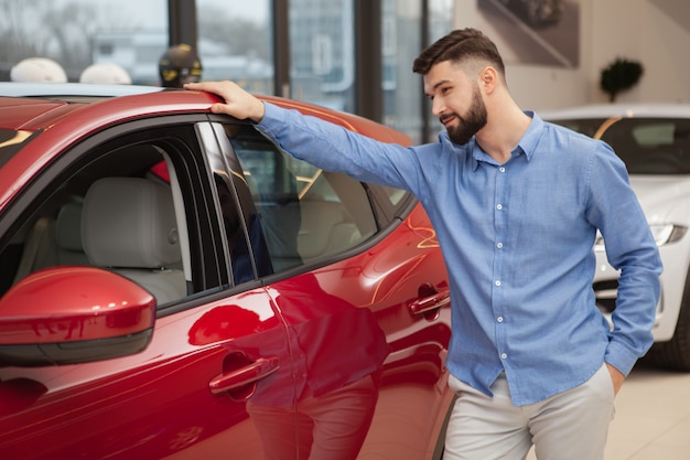 Hombre guapo con barba examinando automóvil rojo en el concesionario de automóviles.