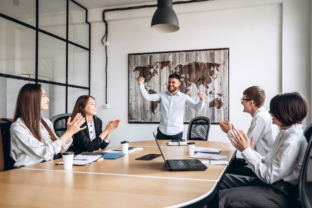 Un hombre guapo con barba feliz joven hace gesto ganador en la oficina de negocios. Se regocija con sus colegas