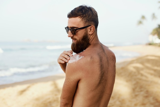 Hombre guapo con barba en gafas de sol tomando el sol con cuerpo de loción de protección solar en verano