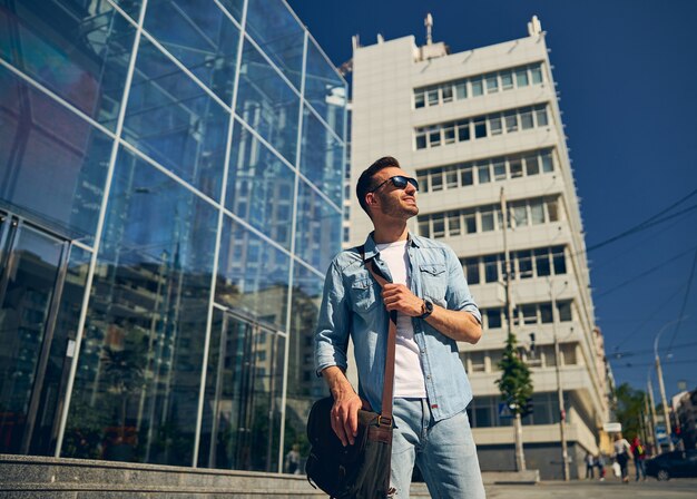 Foto hombre guapo con barba manteniendo una sonrisa en su rostro mientras está de pie cerca del edificio hecho con vidrio