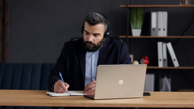 Hombre guapo con barba que estudia en línea usando la cámara web de la computadora portátil, escribiendo una conferencia en el cuaderno.